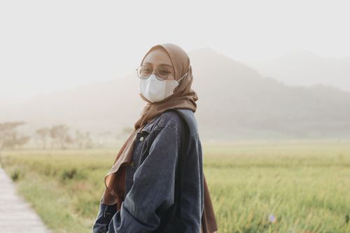 Free A young woman in hijab and mask stands in a serene field in Yogyakarta, Indonesia. Stock Photo
