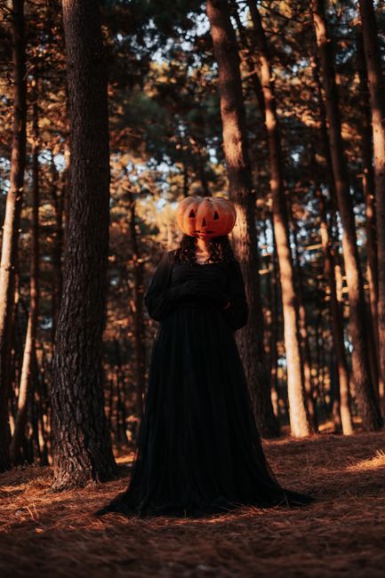 A woman in a black dress with a pumpkin head stands in a forest, evoking a spooky Halloween vibe.