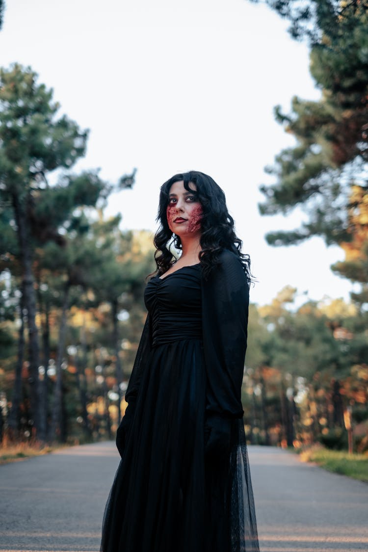 A Woman Wearing A Black Dress With Wound On Her Face Standing Near Trees