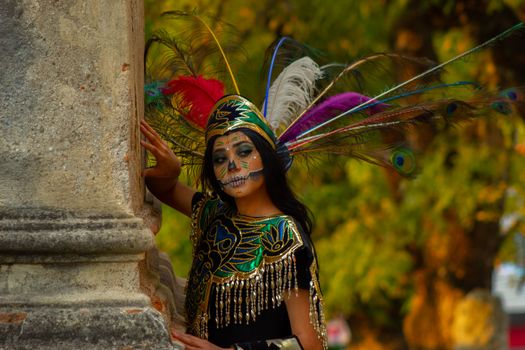 A woman dressed as Catrina with vibrant makeup and feathered headdress in a natural setting.