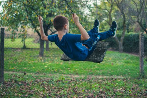 Free A young boy in blue clothes swings joyfully outdoors in a leafy park setting, capturing a moment of childhood bliss. Stock Photo