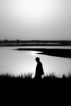 A black and white silhouette of a man walking by the seaside during twilight, capturing a serene moment.
