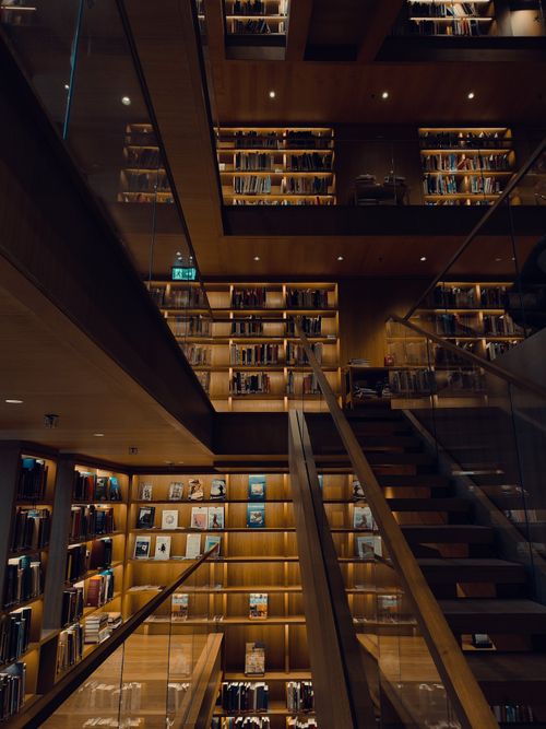 Free Dimly lit modern library with wooden shelves filled with books and glass staircases. Stock Photo
