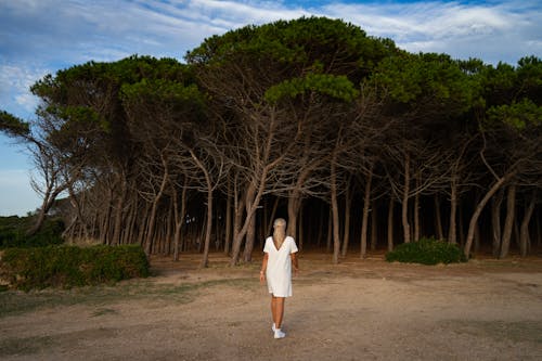 Back View of a Woman in White Dress Walking on a Dirt Ground Near the Trees