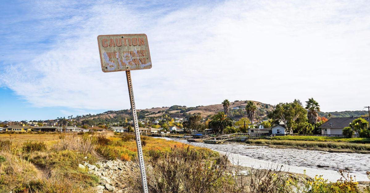 Countryside scene with a weathered caution sign near a river and houses.