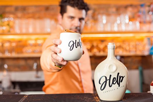 A bartender offers a ceramic mug with 'Help' branding in a modern bar setting, highlighting drink service.