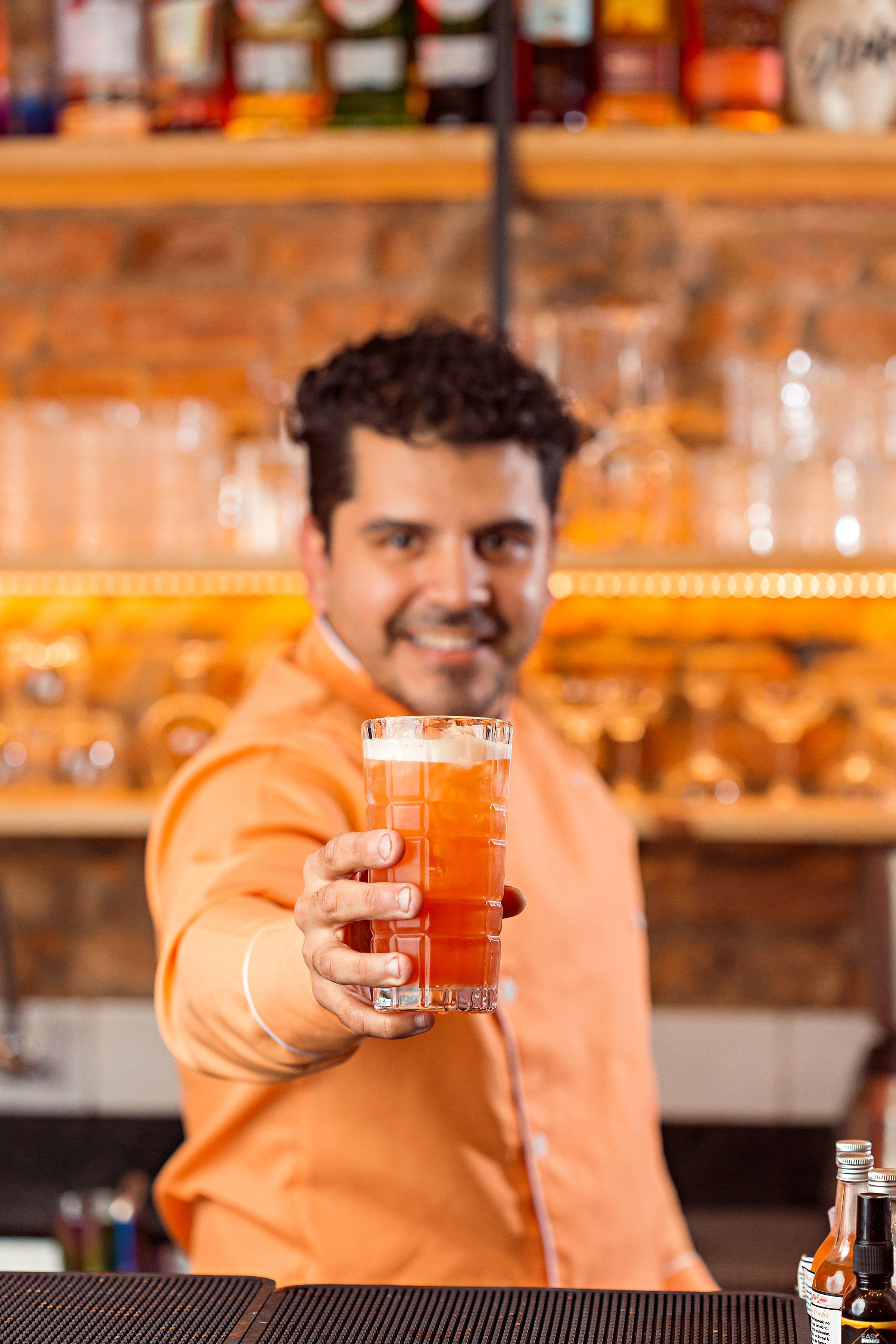 Smiling bartender in orange shirt presenting a refreshing cocktail at a bar.