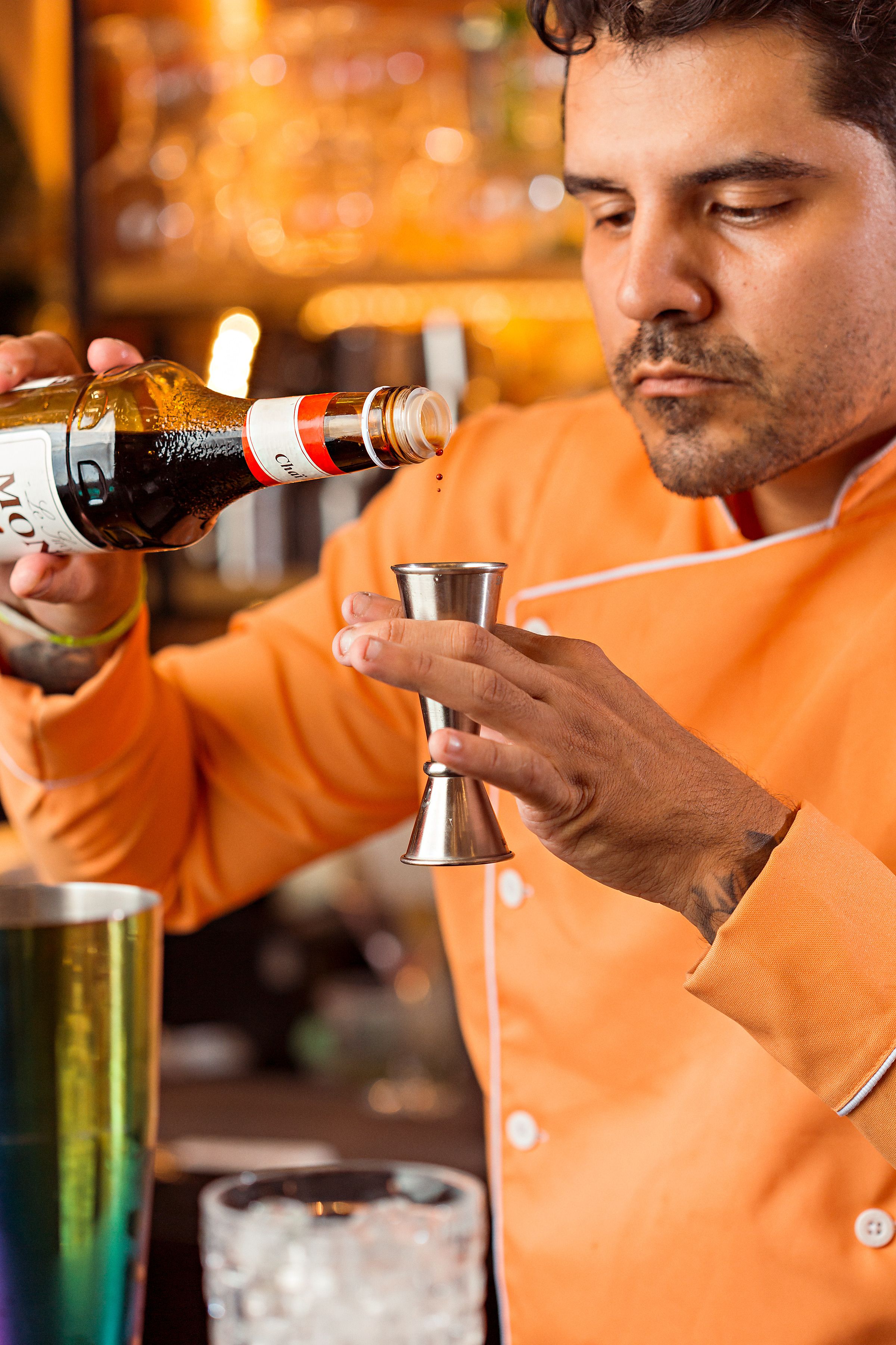 Bartender in orange uniform expertly preparing cocktail with precision pouring in a bar setting.