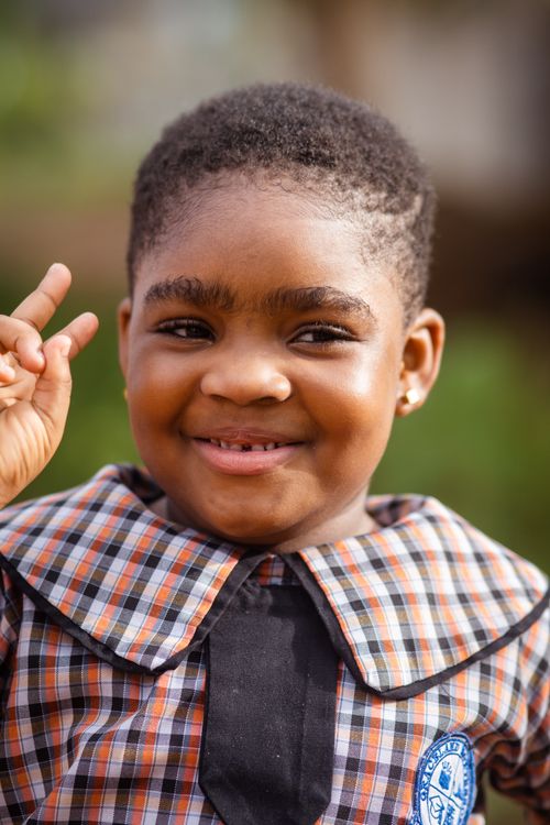 Free A cheerful child with afro hair and plaid uniform smiling outdoors, making a peace sign. Stock Photo