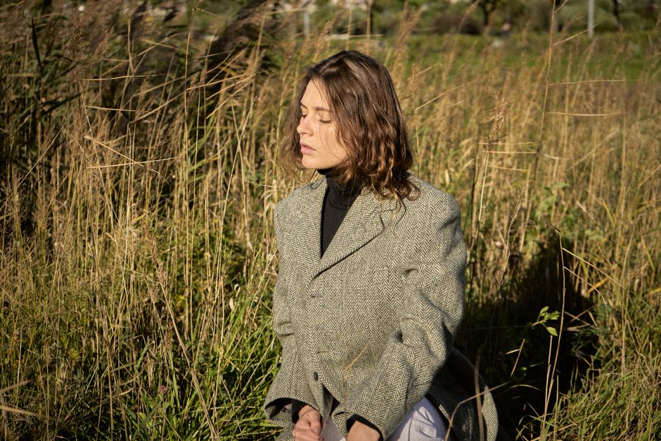 A woman in an oversized olive herringbone tweed blazer over a black turtleneck with light trousers, showing a relaxed modern fit in golden-hour light