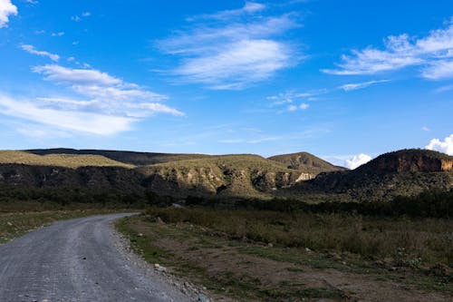 Unpaved Road Between Green Grass Field Near the Mountains Under a Blue Sky