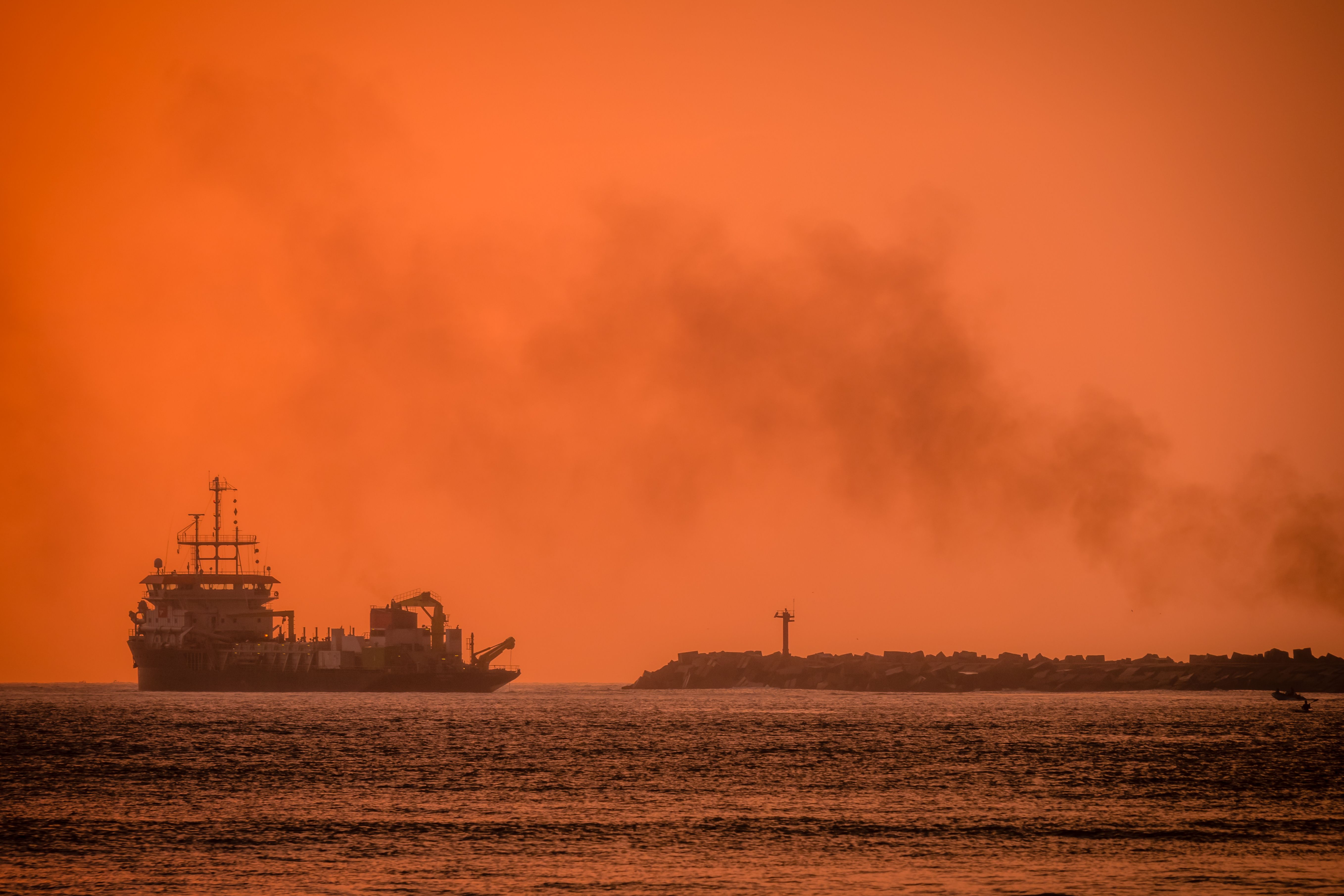 Silhouette of a ship sailing near the coastline during a vibrant orange sunset, emitting smoke against the horizon.