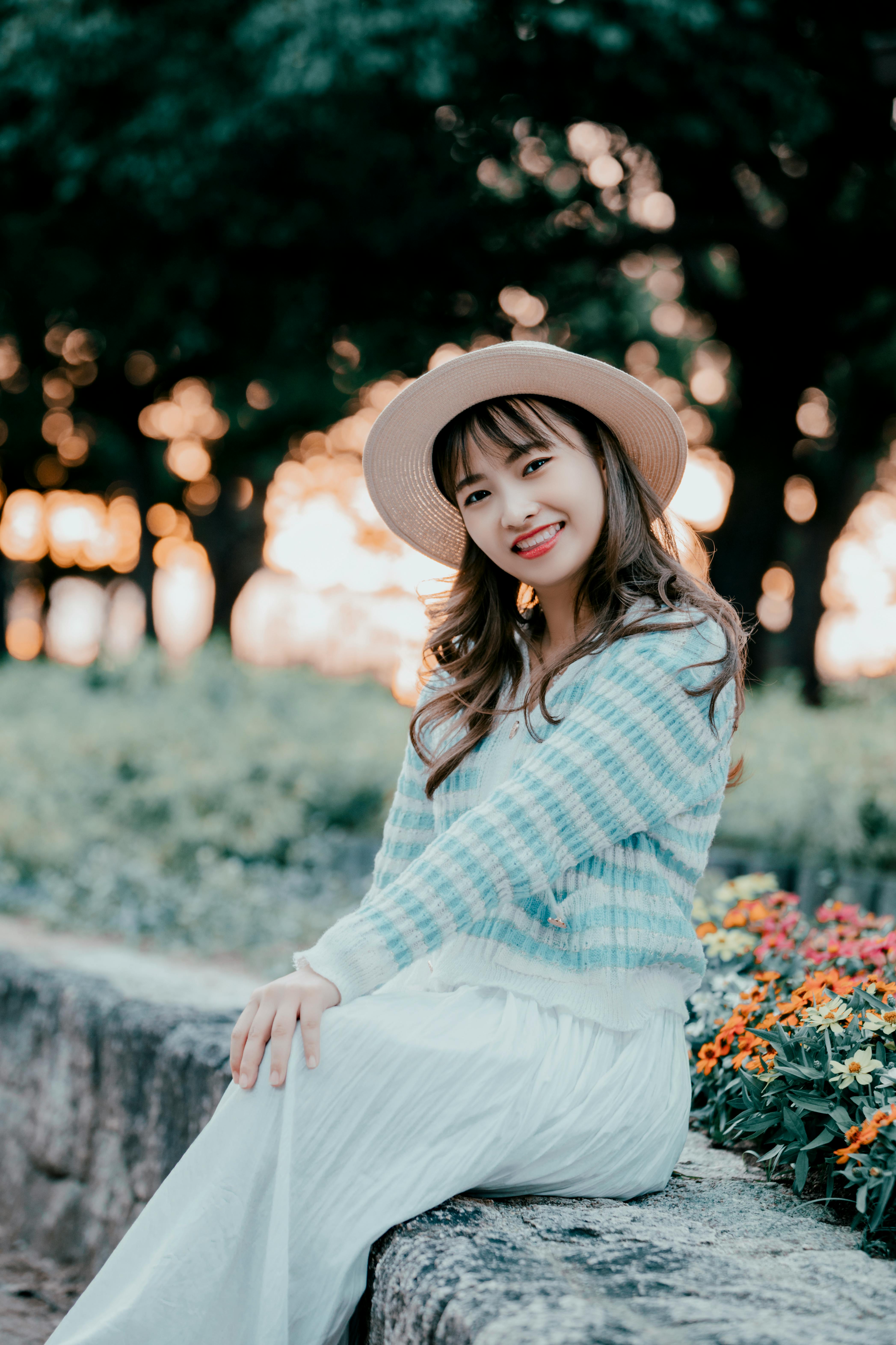 A fashionable Asian woman sits outdoors, smiling in a striped sweater and hat with a warm sunset glo