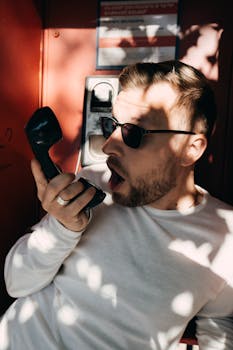 A man wearing sunglasses talks on a phone in a sunlit booth, showcasing urban style in Yerevan, Armenia.