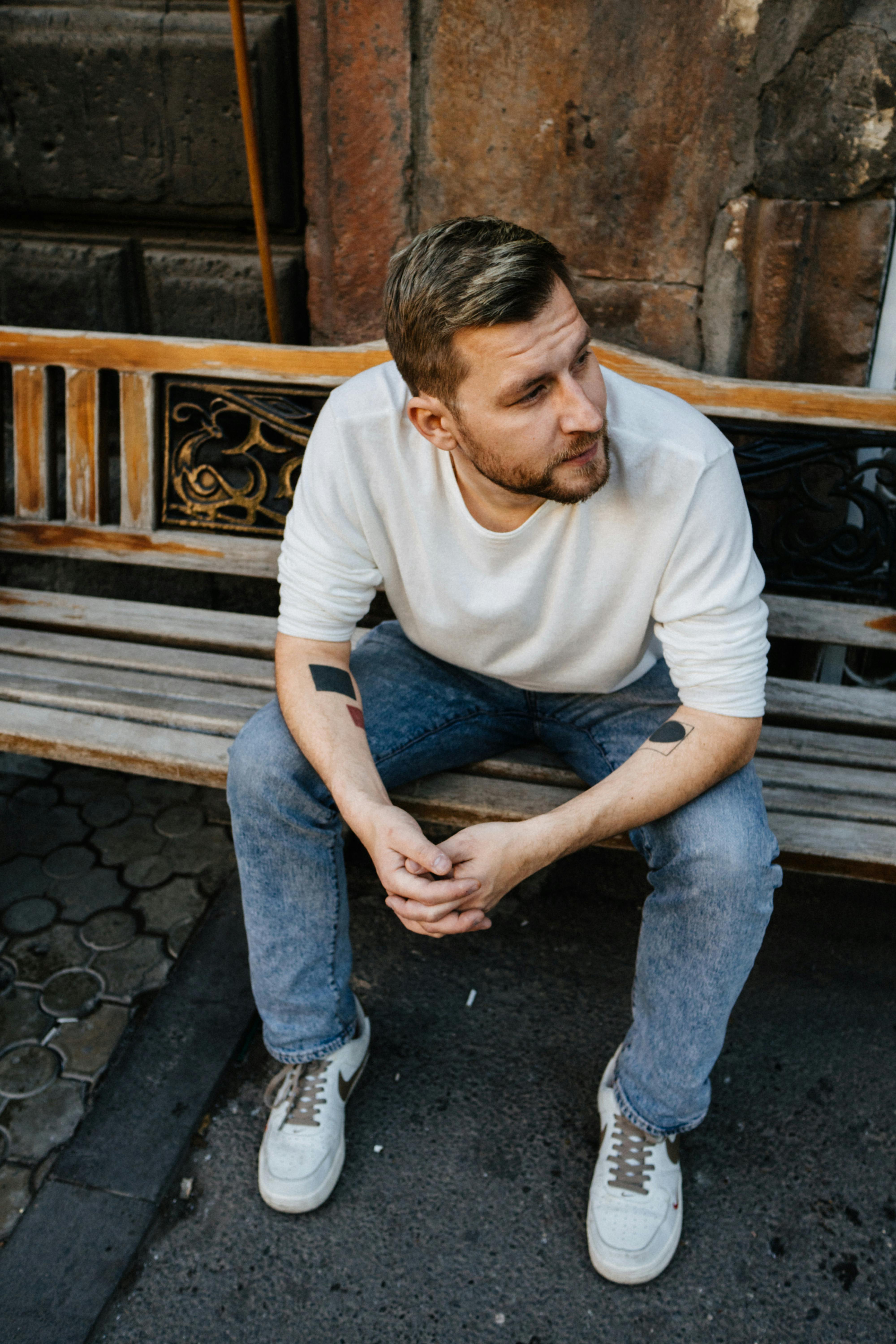 Man with tattoos sitting on a wooden bench in Yerevan, Armenia's urban environment, wearing a white sweater.