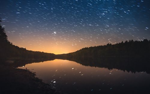 Free A mesmerizing night sky with star trails reflecting on a calm lake surrounded by trees. Stock Photo