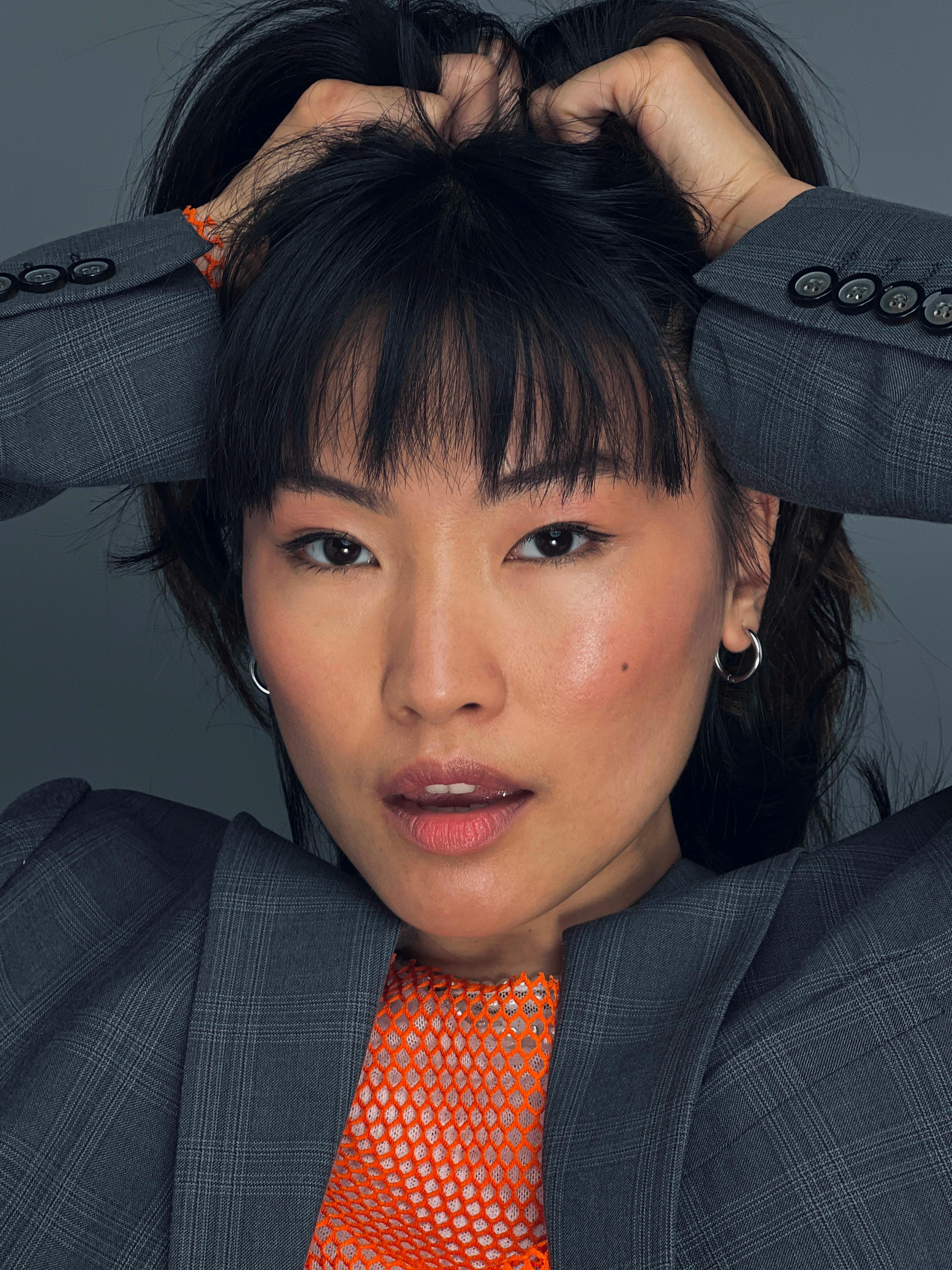 Close-up of a woman in a blazer posing in a studio with her hands in her hair, expressing confidence.