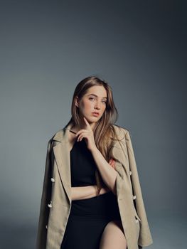 A stylish woman poses in a black dress and beige blazer against a gray studio background.