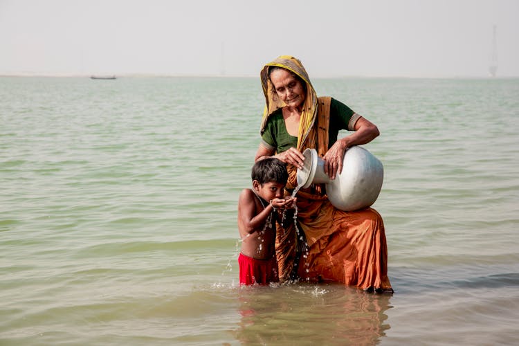 Senior Woman Giving Water From A Vase To A Boy Standing In A River