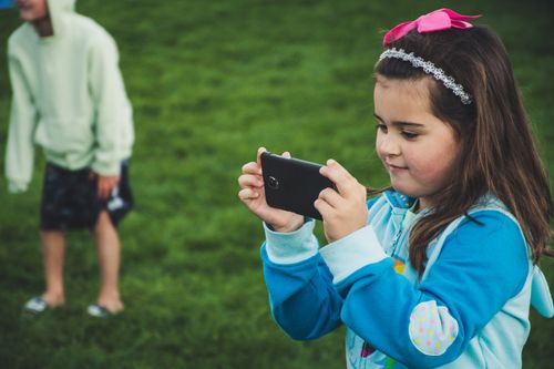 Free A young girl with a smartphone taking a photo in a grassy park setting Stock Photo