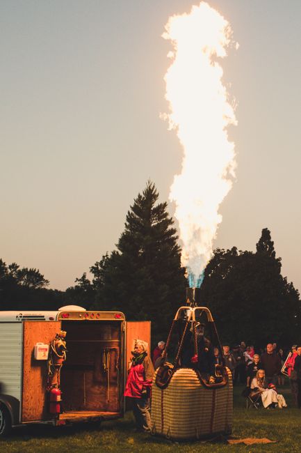 A group prepares a hot air balloon burner for flight at twilight, surrounded by trees and spectators.