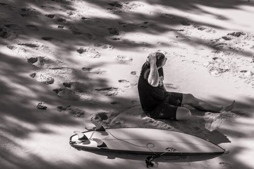 Grayscale Photo of Man Sits Beside Surfboard on Sand