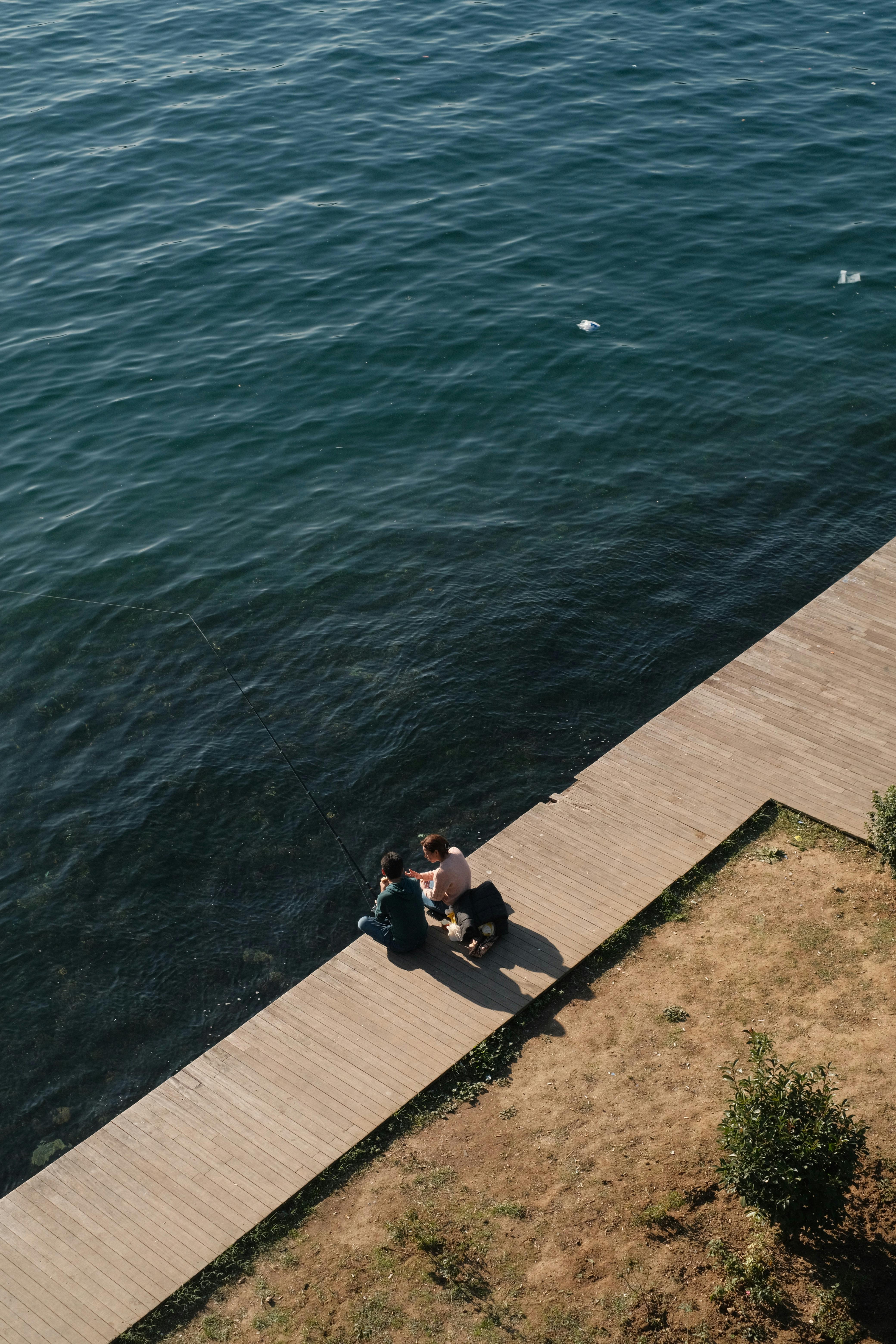High angle view of two people sitting on a boardwalk by the sea, offering a peaceful and relaxed scene.