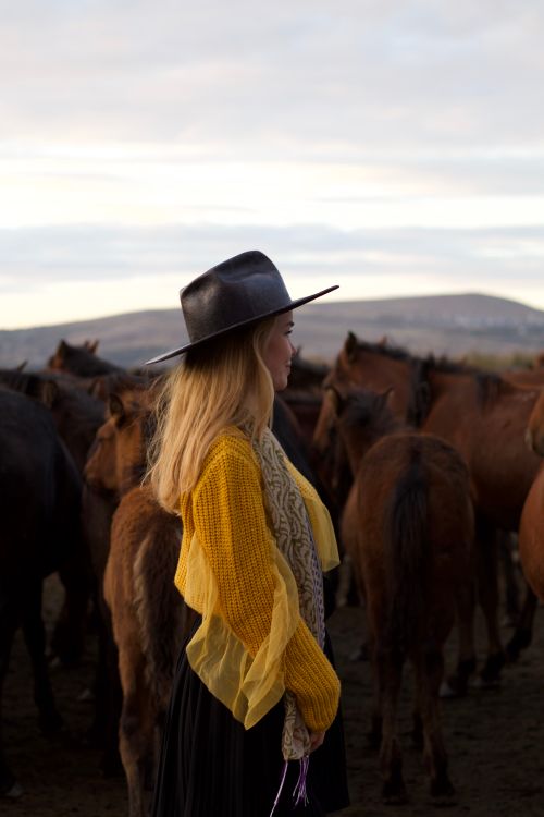 Free Blonde woman with long hair and cowboy hat standing among a herd of horses in a rural setting. Stock Photo