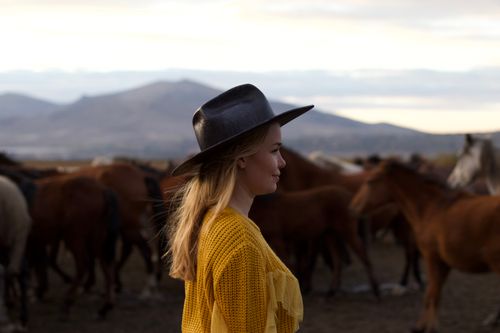Free A woman in a cowboy hat and yellow top stands with horses in a scenic countryside setting. Stock Photo