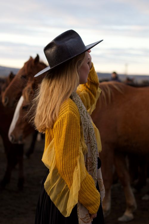 Free Side view of a woman in a yellow sweater and hat standing among horses outdoors at twilight. Stock Photo