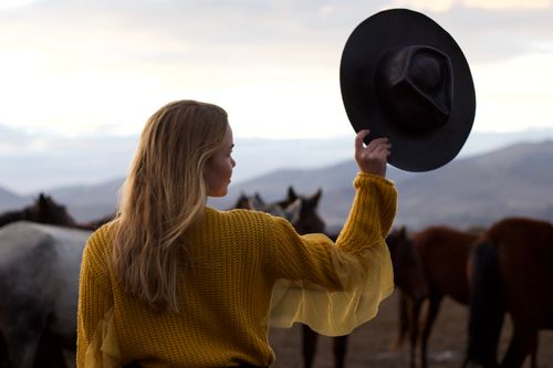 Free Blonde woman in a knitted sweater holding a black hat, standing in a countryside field with horses. Stock Photo