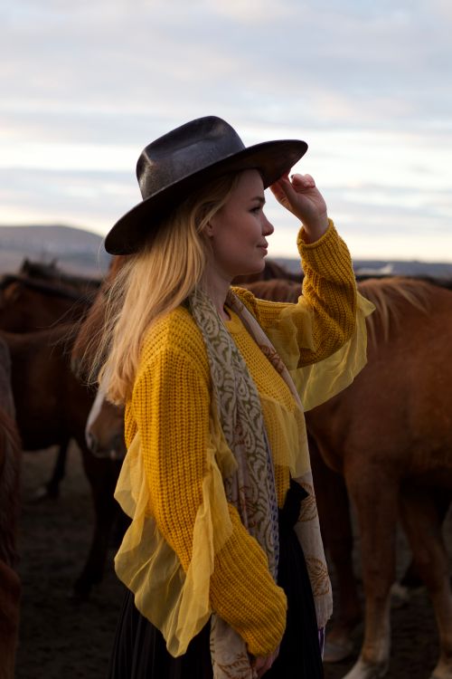 Free Blonde woman in a rural setting with horses, wearing a wide-brim hat, exemplifying cowgirl style. Stock Photo