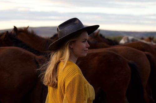 Free Woman in a black hat and yellow sweater standing among horses in a scenic outdoor setting. Stock Photo