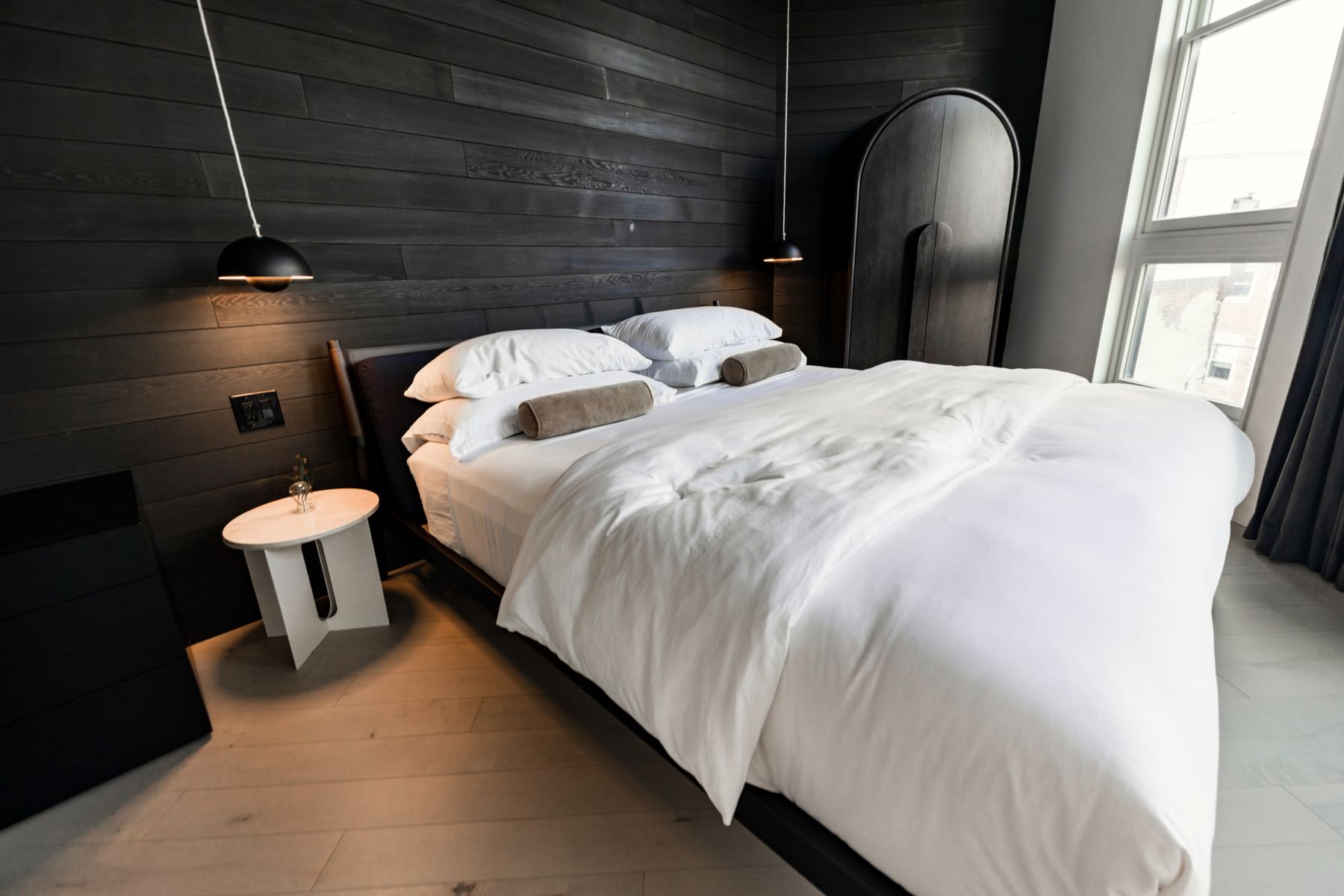 Bedroom wrapped in black vertical wood panels with a pale upholstered bed, white bedding, warm wood sideboard and two dark chairs
