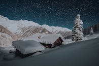 在一个宁静的冬夜，小屋被积雪覆盖，广阔的星空下是白雪皑皑的山脉