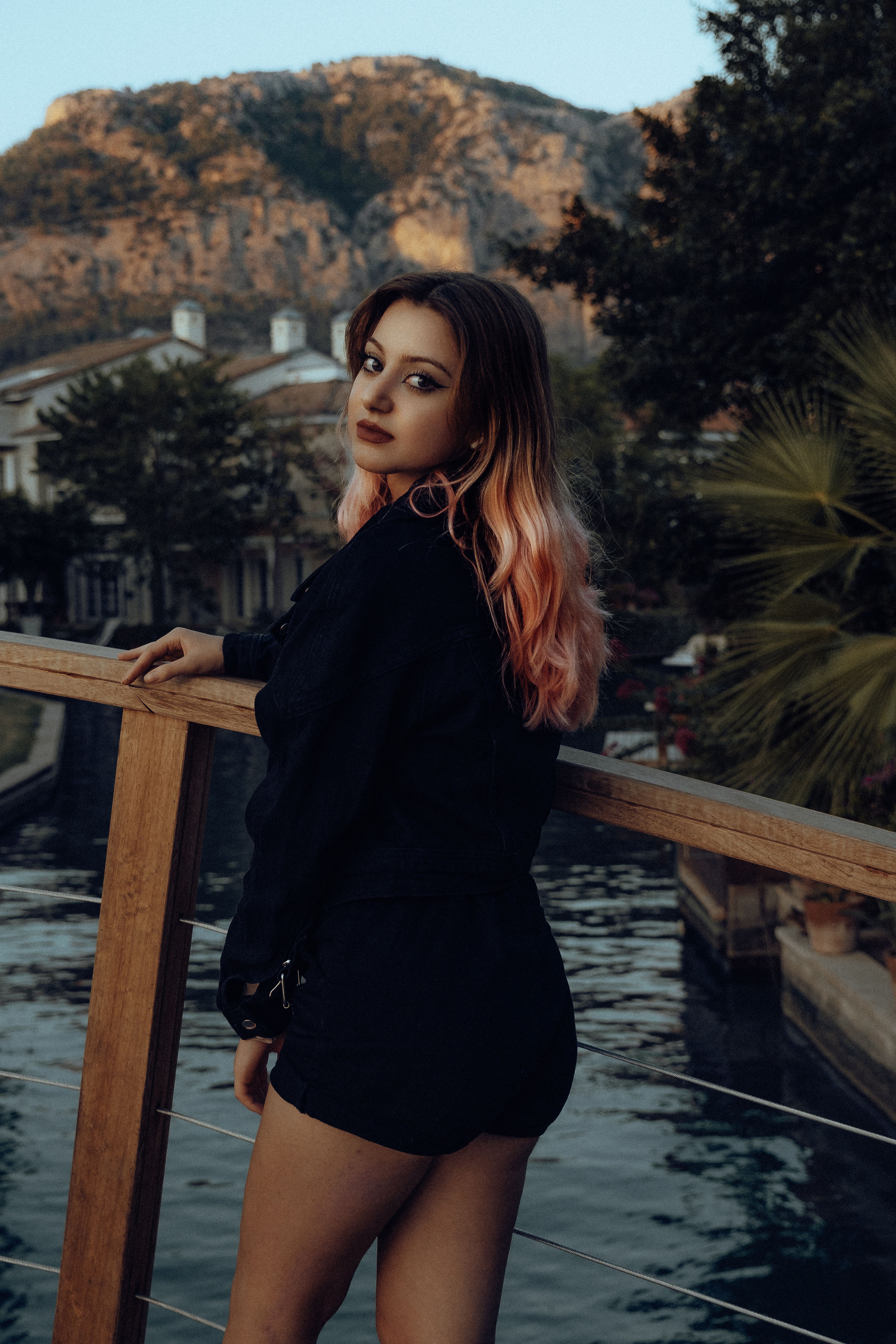 Young woman in black attire stands by railing overlooking a serene landscape with mountains.