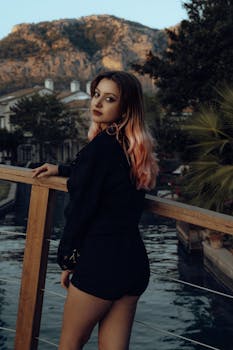 Young woman in black attire stands by railing overlooking a serene landscape with mountains.