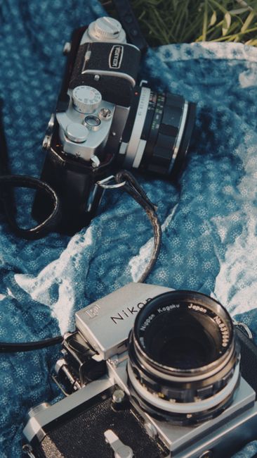 Two vintage film cameras on a blue patterned blanket outdoors during daylight.