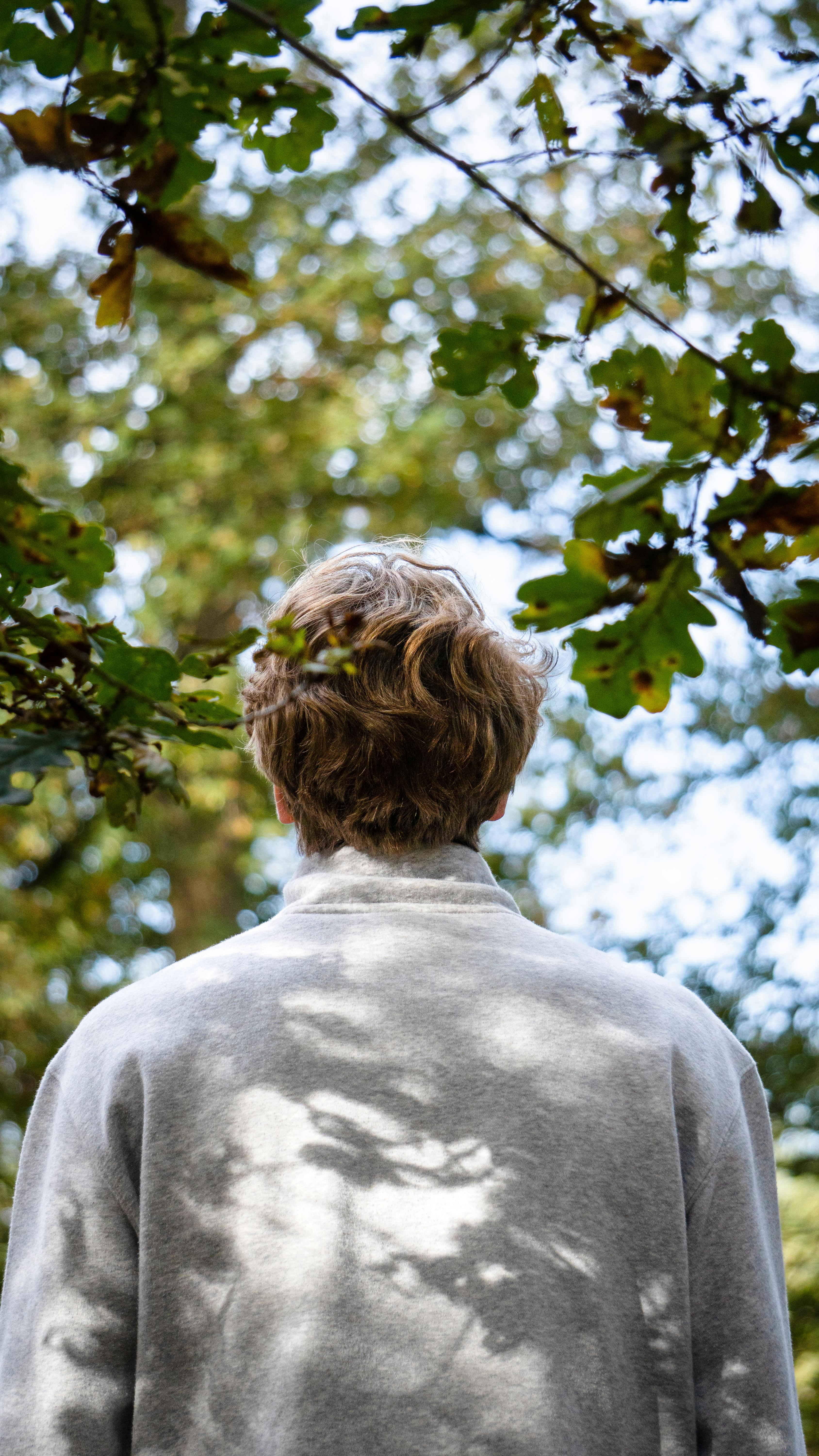 A man with blond hair and white sleeves stands outdoors under a canopy of green leaves, creating a serene nature scene.