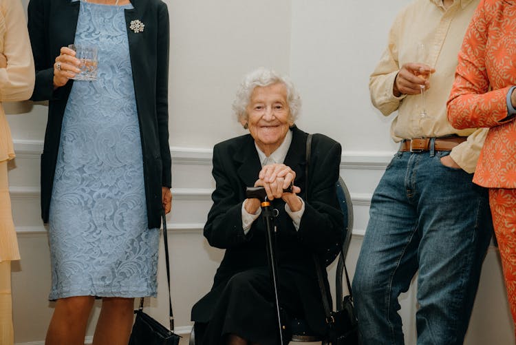 Elderly Woman Sitting At A Party Among Elegantly Dressed People 