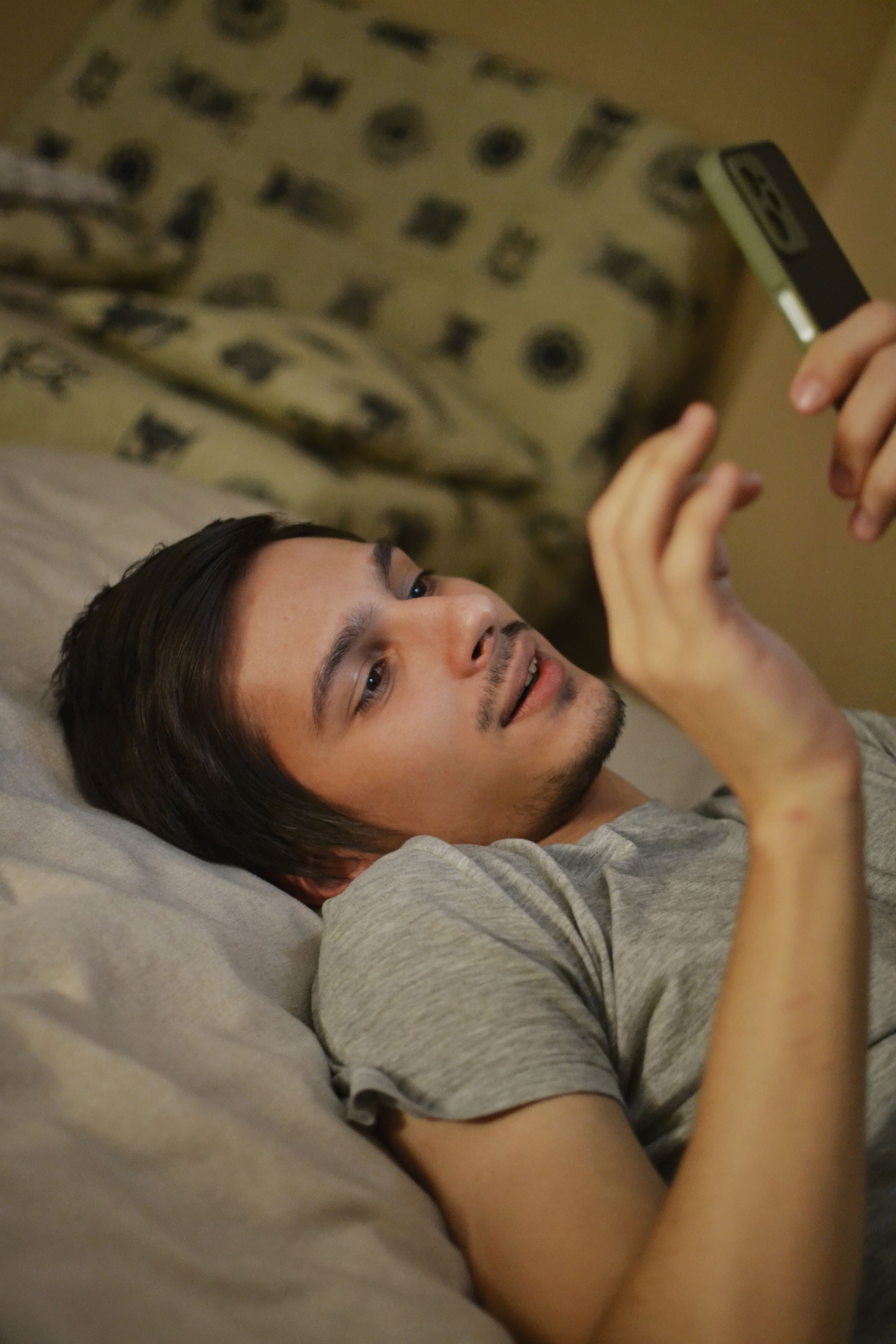 A young adult man relaxes in bed, using a smartphone for leisure.