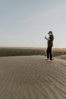 A man standing on a sand dune captures the expansive desert view with his smartphone.