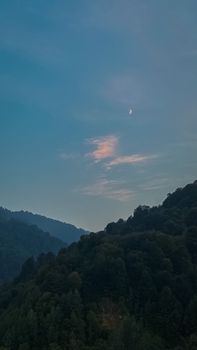 Breathtaking view of hills and a crescent moon in the clear sky during twilight.