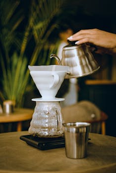 Hand pouring hot water over coffee grounds using a pour-over kettle in a café with plants.