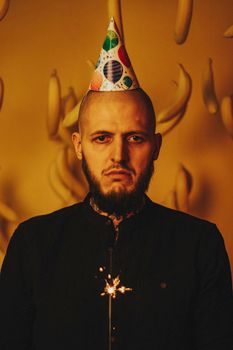 A serious bearded man wearing a party hat holds a sparkler against a yellow background.