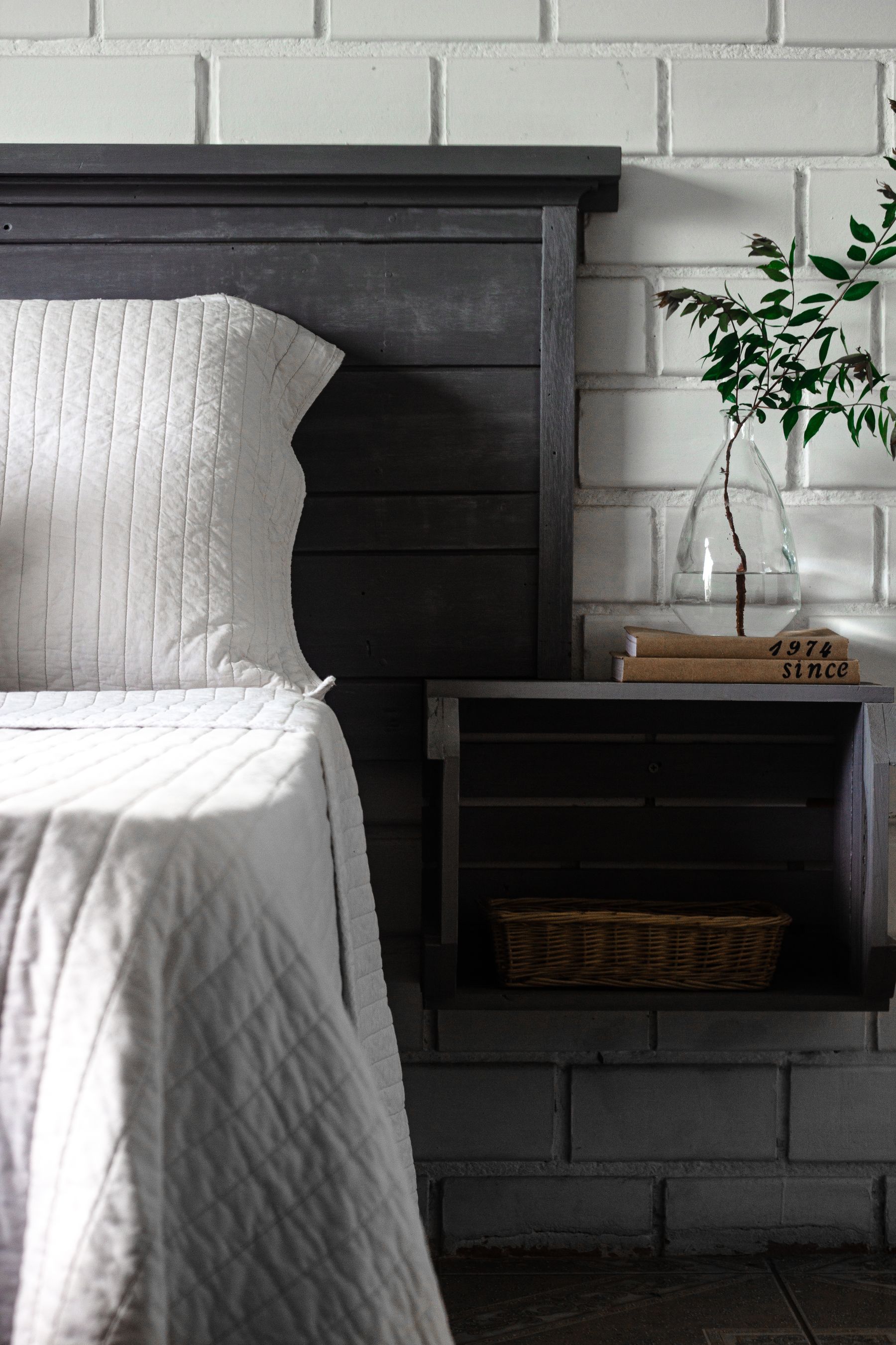 Dark gray timber headboard with pale bedding, a white brick wall and bedside books and vase