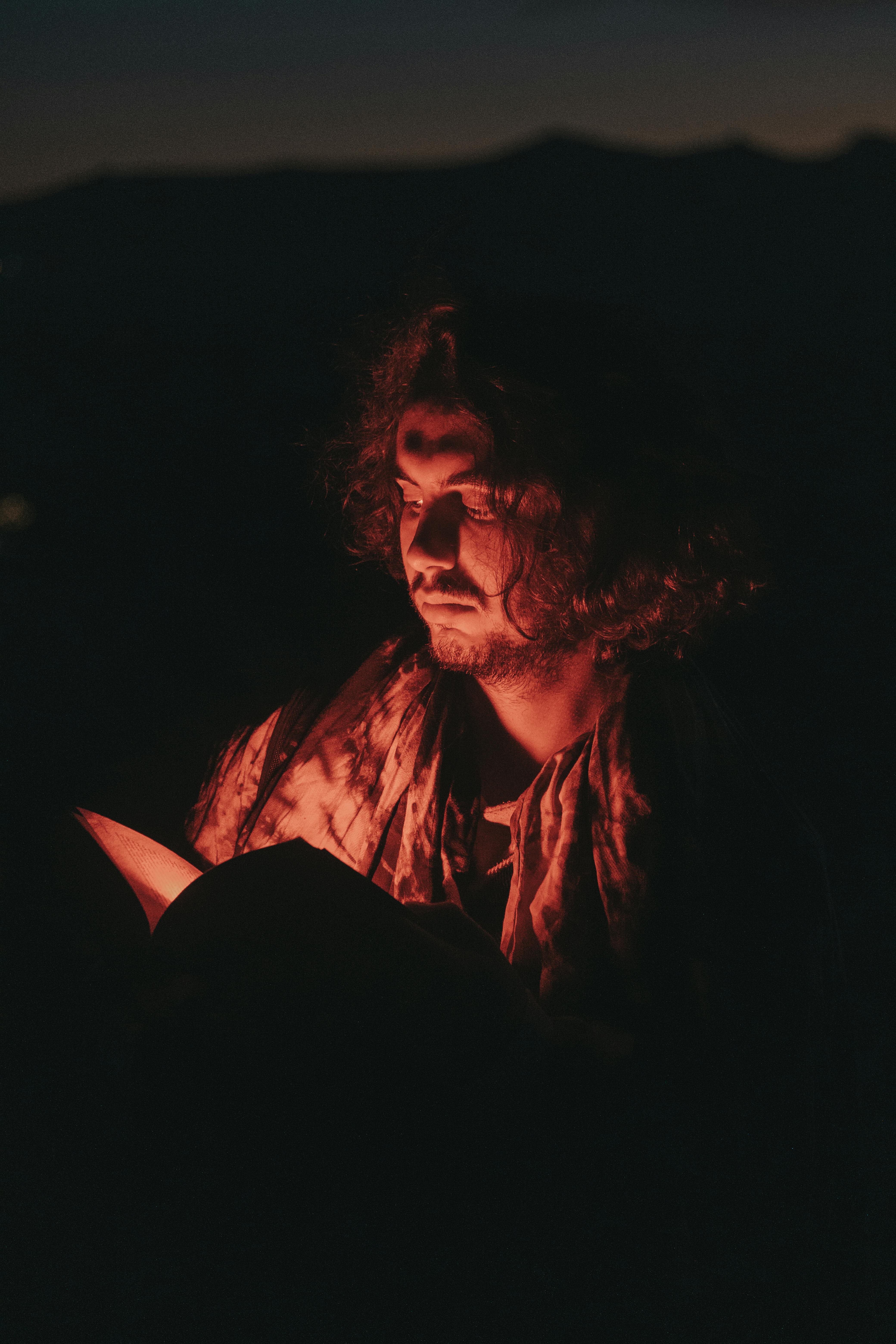 A man reads a book by lamplight casting dramatic shadows on his face at night.