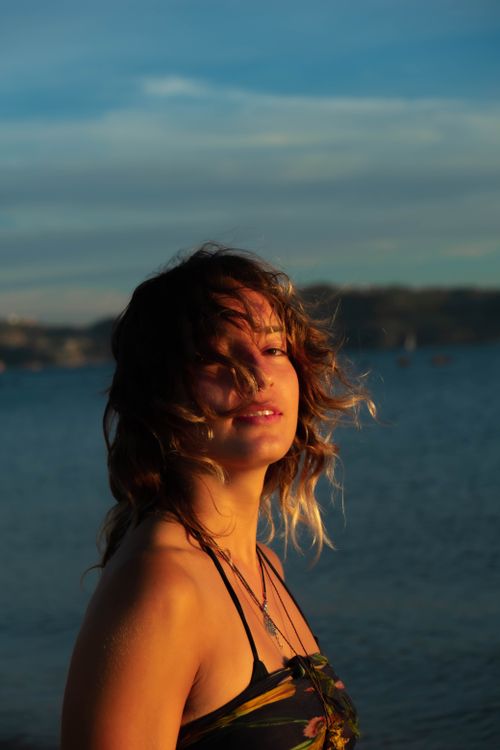 Free A beautiful woman with flowing hair poses by the sea during sunset, creating a serene and dreamy scene. Stock Photo