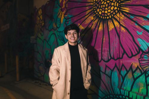 Free Young man in a beige coat smiles in front of a vibrant urban mural at night. Stock Photo