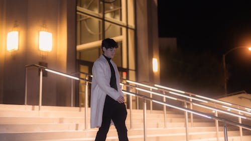 Young Man in a White Coat and Black Turtleneck Walking Down the Stairs in Front of a Building at Night